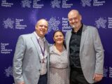 A man in a grey jacket and white shirt, a woman in a blue and white patterned dress and a man in a grey jacket and black shirt stand in front of a purple backdrop featuring the logo and name Forget Me Not children's hospice, at the Race Across West Yorkshire awards evening